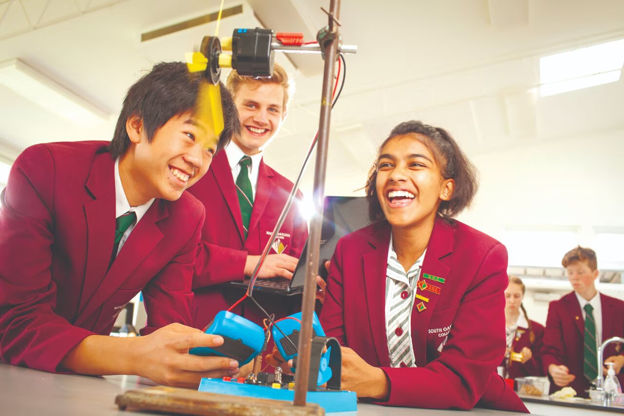 Three high schoolers (two male, one female) smiling as they conduct a class experiment. They are wearing magenta blazers and green ties.